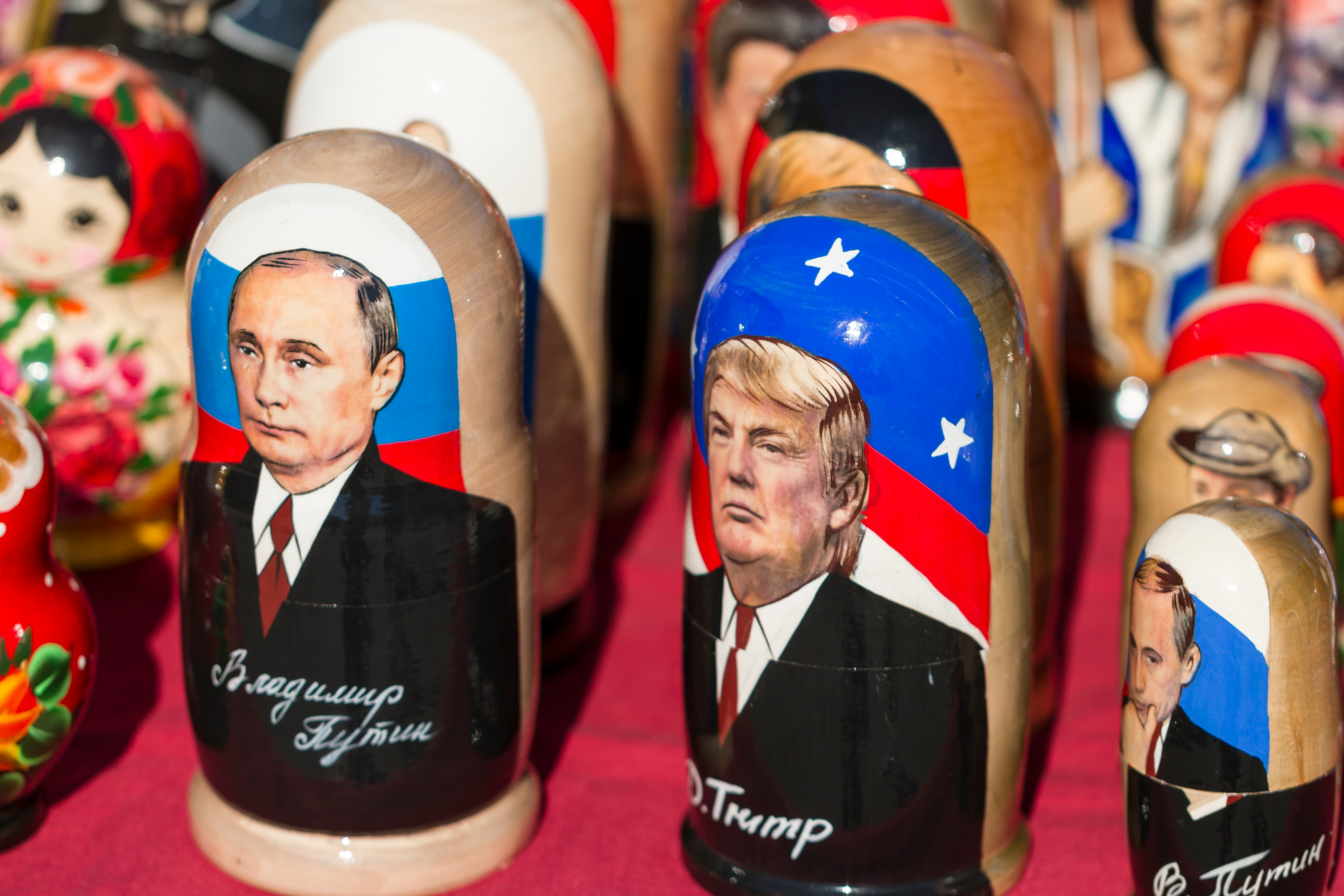 A closeup picture of rows of low-quality Russian nesting dolls at a souvenir stand with its surface covered in red felt. Featured front and center, side by side, are dolls with paintings of Vladimir Putin and Donald Trump. Putin's is labeled "Vladimir Putin" in Cyrillic. Trump's is labeled "D. Trump" in Latin letters. In the background, although it's blurry, you can make out a doll with Elvis Presley on it. Many of the other dolls sport more traditional motifs.