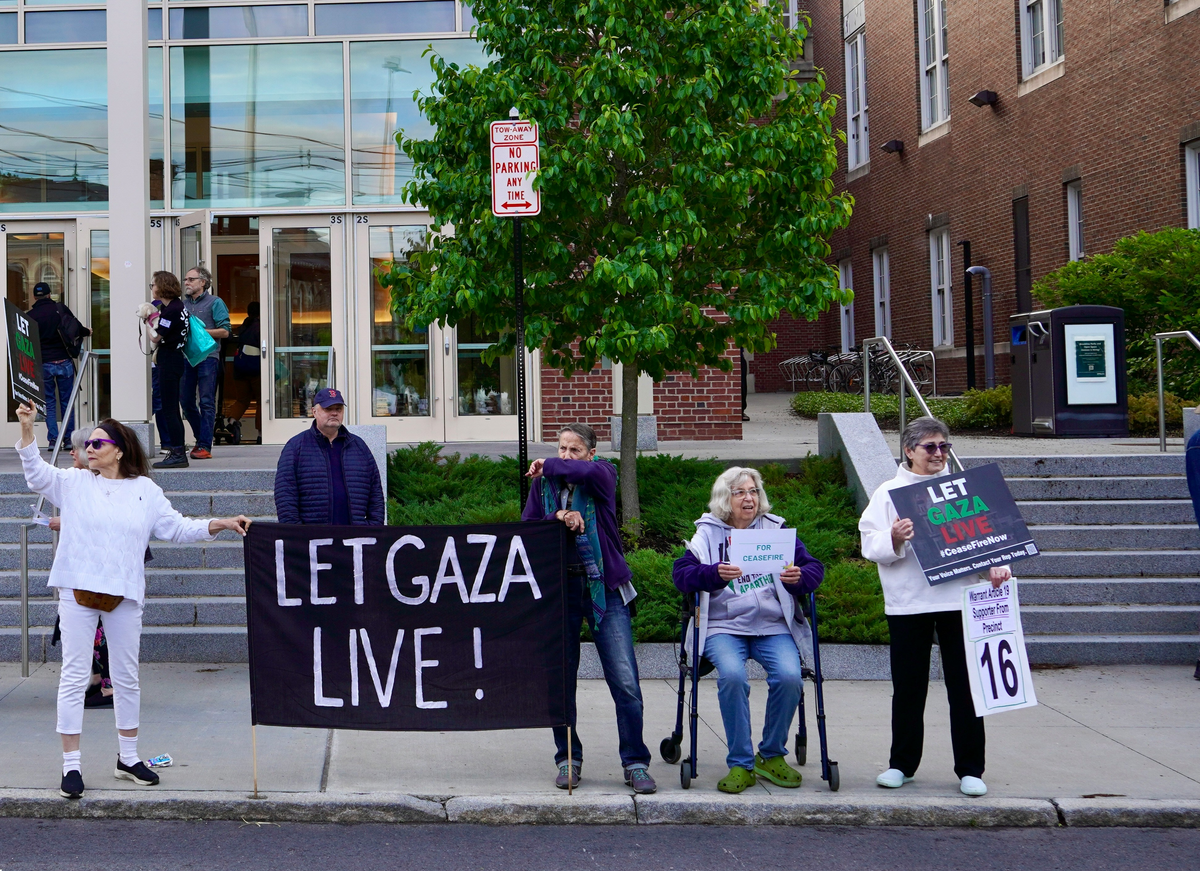 Protesters, most standing and one seated, standing on a sidewalk. Two are holding a large black banner with "LET GAZA LIVE!" on it in large block letters.