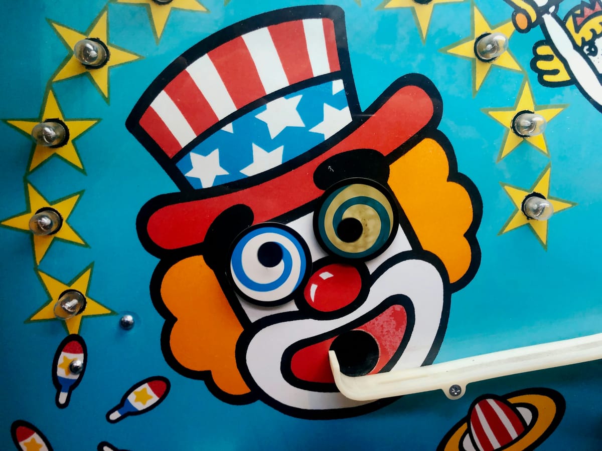 Photo by James Lee, via Unsplash. A picture of a clown-faced, red-nosed, Uncle Sam like figure with swirling eyes on a blue background. It appears to be a pinball machine.
